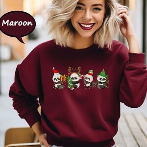 May include: A maroon sweatshirt featuring a festive design of three pandas wearing Santa hats and holding baubles. The pandas are surrounded by Christmas lights and foliage.