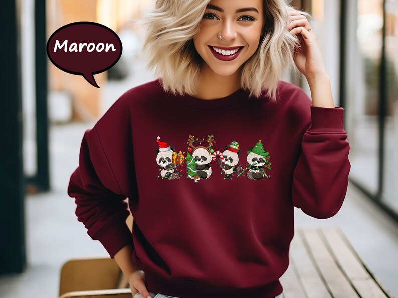 May include: A maroon sweatshirt featuring a festive design of three pandas wearing Santa hats and holding baubles. The pandas are surrounded by Christmas lights and foliage.