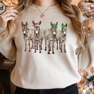 May include: A sand-coloured sweatshirt with a Christmas design. It features four donkeys wearing festive hats, scarves, and fairy lights. The donkeys are arranged in a row. The sweatshirt is a neutral colour, perfect for the winter season.