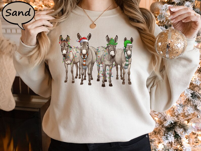 May include: A sand-coloured sweatshirt with a Christmas design. It features four donkeys wearing festive hats, scarves, and fairy lights. The donkeys are arranged in a row. The sweatshirt is a neutral colour, perfect for the winter season.