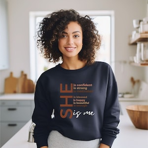 Op de afbeelding: Een marineblauw sweatshirt met de tekst "SHE is confident is strong is motivated is blessed is happy is beautiful is enough is me" in bruin gedrukt op de voorkant.