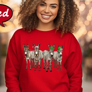 May include: A red sweatshirt with a Christmas design. The design features four donkeys wearing festive hats, scarves, and fairy lights. The word "Red" is in a speech bubble in the upper left corner.