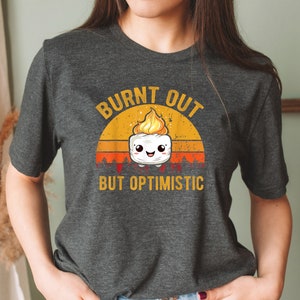 May include: A dark gray t-shirt with a retro-style graphic of a burnt marshmallow with the text "Burnt Out But Optimistic".
