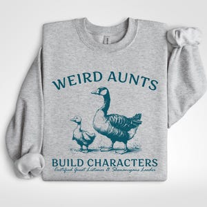 May include: A light gray sweatshirt with the words "WEIRD AUNTS" in teal above a teal illustration of a goose and a gosling. Below the illustration, the text reads "BUILD CHARACTERS" and "Certified Great Listener & Shenanigans Leader."