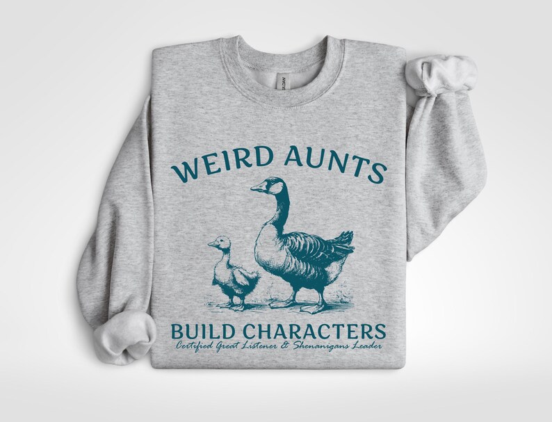 May include: A light gray sweatshirt with the words "WEIRD AUNTS" in teal above a teal illustration of a goose and a gosling. Below the illustration, the text reads "BUILD CHARACTERS" and "Certified Great Listener & Shenanigans Leader."