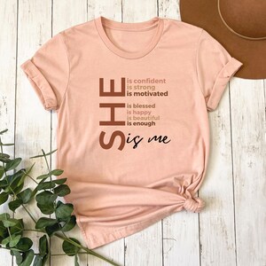 Op de afbeelding: Een roze T-shirt met het woord "SHE" in grote bruine letters. De letters staan verticaal op elkaar en elke letter heeft een andere bruine tint. De tekst "is confident, is strong, is motivated, is blessed, is happy, is beautiful, is enough, is me" staat in kleinere bruine letters naast de letters van het woord "SHE".
