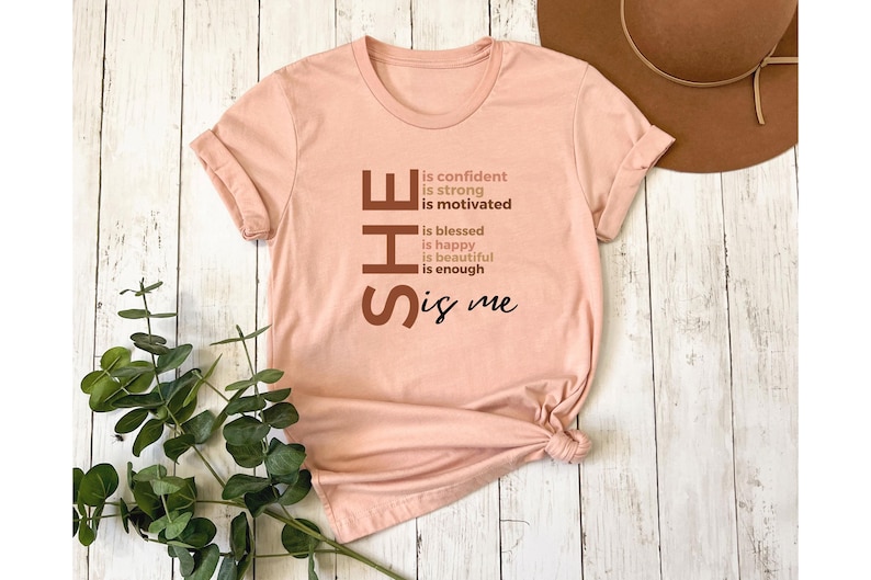 Op de afbeelding: Een roze T-shirt met het woord "SHE" in grote bruine letters. De letters staan verticaal op elkaar en elke letter heeft een andere bruine tint. De tekst "is confident, is strong, is motivated, is blessed, is happy, is beautiful, is enough, is me" staat in kleinere bruine letters naast de letters van het woord "SHE".