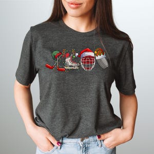 May include: A dark heather grey t-shirt with a Christmas-themed hockey design. The design features a hockey stick, skates, a hockey helmet, and a puck, all decorated with Christmas lights and Santa hats.