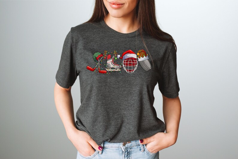 May include: A dark heather grey t-shirt with a Christmas-themed hockey design. The design features a hockey stick, skates, a hockey helmet, and a puck, all decorated with Christmas lights and Santa hats.