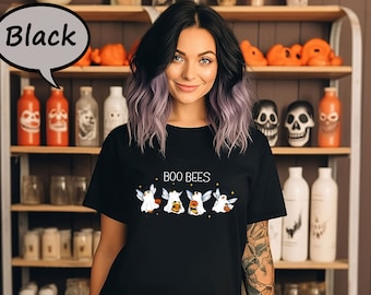 May include: Black t-shirt with the text "BOO BEES" and a graphic of four cartoon bees dressed as ghosts, with wings and holding pumpkins. The shirt is displayed on a person in front of a shelf with Halloween decorations.