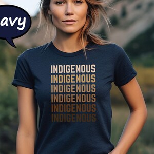 Pu&ograve; includere: T-shirt blu navy con la parola "INDIGENOUS" stampata in una sfumatura di toni oro, ripetuta sei volte. La maglietta &egrave; a girocollo. Una nuvoletta blu navy con la parola "Navy" si trova nell'angolo in alto a sinistra.