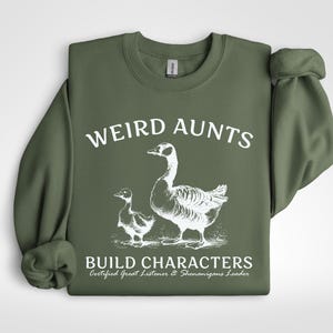 May include: An olive green sweatshirt with the words "WEIRD AUNTS" in white block letters above a white illustration of a goose and a gosling. Below the illustration, the text reads "BUILD CHARACTERS" and "Certified Great Listener & Shenanigans Leader."