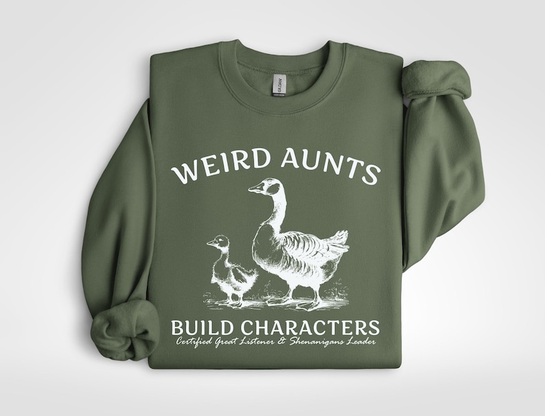 May include: An olive green sweatshirt with the words "WEIRD AUNTS" in white block letters above a white illustration of a goose and a gosling. Below the illustration, the text reads "BUILD CHARACTERS" and "Certified Great Listener & Shenanigans Leader."