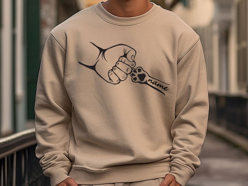 Pu&ograve; includere: Una felpa beige con una grafica nera di una mano umana e un'impronta di zampa che si stringono la mano. Il testo "name" &egrave; scritto sotto l'impronta di zampa.
