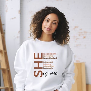 Op de afbeelding: Witte trui met de tekst "SHE is confident is strong is motivated is blessed is happy is beautiful is enough is me" gedrukt in bruin en roze.