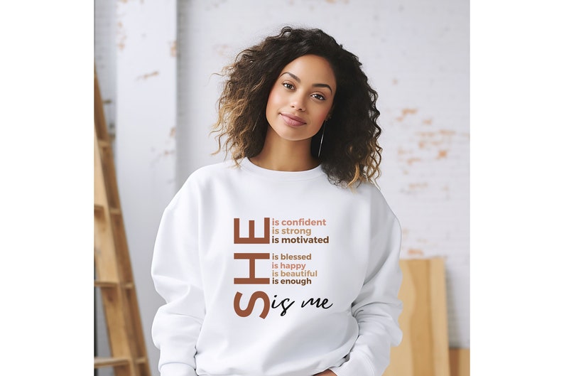 Op de afbeelding: Witte trui met de tekst "SHE is confident is strong is motivated is blessed is happy is beautiful is enough is me" gedrukt in bruin en roze.