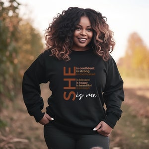 Op de afbeelding: Zwart sweatshirt met de tekst "SHE is confident is strong is motivated is blessed is happy is beautiful is enough is me" gedrukt in bruin en wit.