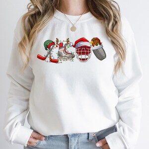 May include: White sweatshirt with a Christmas-themed hockey design. The design features a hockey stick, a hockey skate, a hockey helmet, and a puck, all decorated with Christmas lights and ornaments.