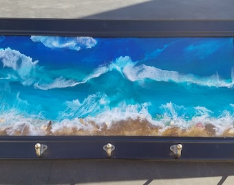 May include: A black framed key holder with a resin ocean scene. The scene features blue water with white foamy waves crashing on a sandy beach. The key holder has four hooks.