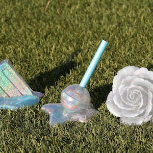 May include: Three iridescent, melted, and glittery objects on a green grassy surface. A popsicle, a lollipop, and a white rose.