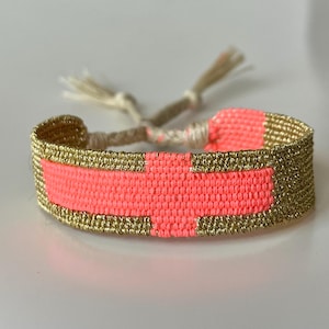 May include: A woven friendship bracelet with a pink and gold design. The bracelet has a white cord with tassels.