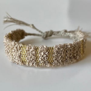 May include: A beige and gold braided bracelet with a tassel. The bracelet is made of a soft, textured material and has a simple, elegant design.