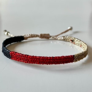 Può includere: Braccialetto intrecciato con un design a blocchi di colore. Il braccialetto presenta un motivo nero, rosso e beige. Il braccialetto regolabile ha una chiusura color argento e un cordino beige.