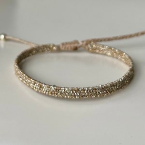 Könnte beinhalten: Ein geflochtenes Armband in Gold und Beige mit einem verschiebbaren Knotenverschluss.
