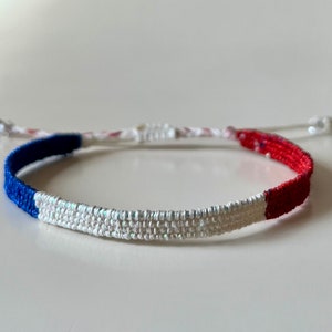 Könnte beinhalten: Ein gewebtes Freundschaftsarmband mit blauen, weißen und roten Abschnitten. Das Armband hat einen Schiebeknotenverschluss und besteht aus gewebten Fäden. Die Farben sind in einem Streifenmuster angeordnet, mit Blau und Rot an den Enden.