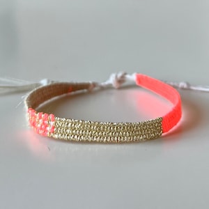 Könnte beinhalten: Ein goldfarbenes und korallenfarbenes Armband mit einer weißen Schnur und einem Quastenanhänger. Das Armband besteht aus kleinen, runden Perlen und hat ein schlichtes, elegantes Design.