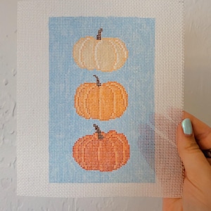 May include: A needlepoint canvas featuring a vertical arrangement of three pumpkins in varying shades of cream, light orange, and dark orange, all set against a light blue rectangular background on a white mesh grid.