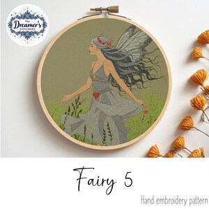 May include: An embroidery hoop featuring a fairy design. The fairy is depicted in a flowing gray dress with gray wings and long gray hair. The background is a muted green with embroidered grass and flowers. The text "Fairy 5" is below the hoop.