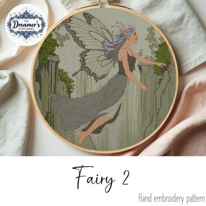 May include: Hand embroidery pattern of a fairy with butterfly wings, wearing a gray dress, and a floral crown. The fairy is set against a green and gray background, with the text "Fairy 2" and "Hand embroidery pattern". The embroidery is in a wooden hoop.