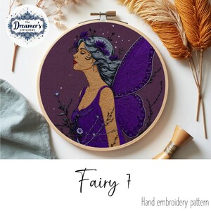 Peut inclure: Broderie à la main représentant une fée violette avec de grandes ailes et une robe violette, dans un cercle en bois. Le texte "Fairy 7" et "Hand embroidery pattern" sont visibles.