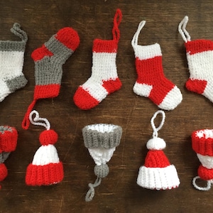 Può includere: Dieci ornamenti natalizi fatti a mano, tra cui calze e cappelli, in rosso, bianco e grigio. Gli ornamenti sono all'uncinetto e hanno un tema natalizio festivo.