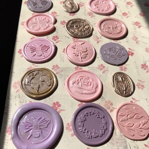 May include: Assortment of wax seals in various colors and designs, including bees, flowers, and wreaths. The seals are arranged on a patterned surface.