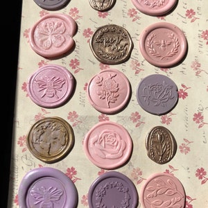 May include: A collection of 13 wax seals with various designs, including bees, flowers, leaves, and a heart with the text "Forever Yours". The seals are in shades of pink, purple, and gold.