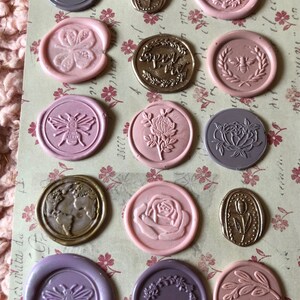 May include: Assortment of 13 wax seals with various designs, including bees, flowers, leaves, and a globe. The seals are in shades of pink, purple, and gold.
