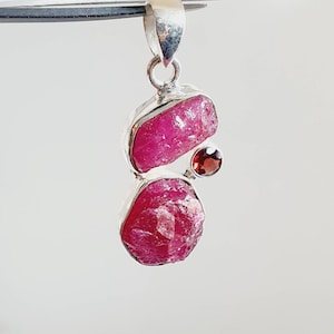 Peut inclure: Un pendentif en argent avec deux pierres de rubis brutes et une plus petite pierre de grenat. Les pierres de rubis sont d'une couleur rose foncé et ont une texture brute et naturelle. Le pendentif est un bijou unique et accrocheur.