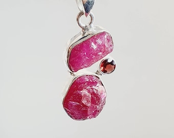 May include: A silver pendant featuring two raw ruby stones and a smaller garnet stone. The ruby stones are a deep pink color and have a rough, natural texture. The pendant is a unique and eye-catching piece of jewelry.