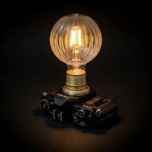 Può includere: Una lampada unica realizzata con un corpo macchina Canon vintage. La base nera della fotocamera supporta una lampadina decorativa in vetro a coste con un filamento caldo e luminoso. La lampada è ambientata su uno sfondo scuro.
