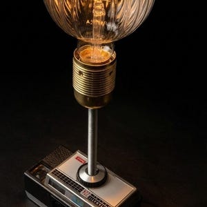 Peut inclure: Lampe originale avec une base de caméra Kodak Instamatic 224 vintage. La lampe a une douille de lampe de couleur laiton et une grande ampoule en verre clair et nervuré. L'ampoule est allumée, émettant une lueur chaleureuse. La lampe est un mélange créatif de rétro et de moderne.