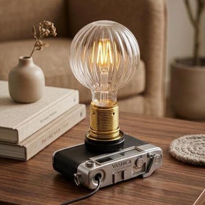 Könnte beinhalten: Eine Tischleuchte mit einem silber-schwarzen Vintage-Kameragehäuse von Yashica als Basis, gekrönt von einer gerippten, klaren Glaskugel-Glühbirne. Die Lampe hat eine messingfarbene Fassung und ein schwarzes Stromkabel an der Vorderseite.