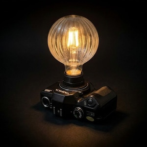 Könnte beinhalten: Eine einzigartige Schreibtischlampe mit einem Vintage-Yashica-Kameragehäuse und einer klaren, gerippten Glühbirne. Das schwarze Kameragehäuse hat silberne Akzente und Knöpfe. Die Glühbirne strahlt ein warmes Licht vor dunklem Hintergrund aus.