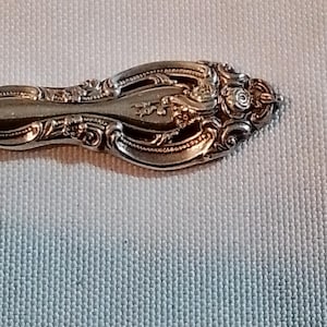 May include: Close-up of a silver spoon with ornate floral and scrollwork detailing. The spoon's handle features a raised design with a rose at the end. The spoon is set against a light-colored, textured background.