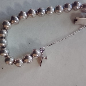 May include: A silver-toned beaded bracelet composed of smooth, spherical metallic beads strung together. The bracelet features a rectangular box clasp and a thin safety chain attached to a small, screw-back style metal component.