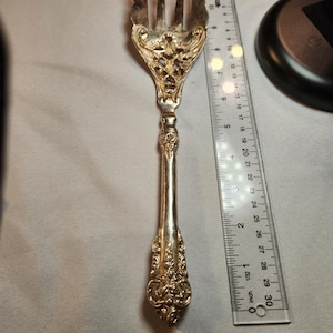 May include: A silver ornate serving fork with a decorative handle and three tines. The fork is approximately 8 inches long.