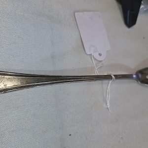 May include: A silver-toned metal cocktail fork with a long, slender handle featuring a decorative border and an engraved script letter. The fork head has three tines and a small central opening. A white paper tag is attached with a thin white string.