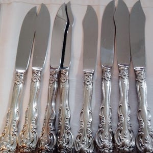 May include: A set of eight silver butter knives. The knives have a curved blade and ornate, detailed handles with a floral design. The blades are shiny and reflect the light, while the handles have a matte finish. The knives are arranged on a white surface.