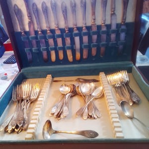 May include: A silver-toned cutlery set housed in a wooden storage case with a green velvet interior. The set includes knives arranged in the lid, along with various forks, spoons, and a serving ladle organised in the base compartments.
